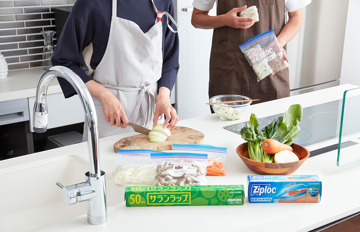 ヘーベルハウス馬込展示場のキッチンで冷凍貯金をするために野菜を切る女性と、ジップロックにおにぎりを入れようとする男性。サランラップとジップロックのパッケージ